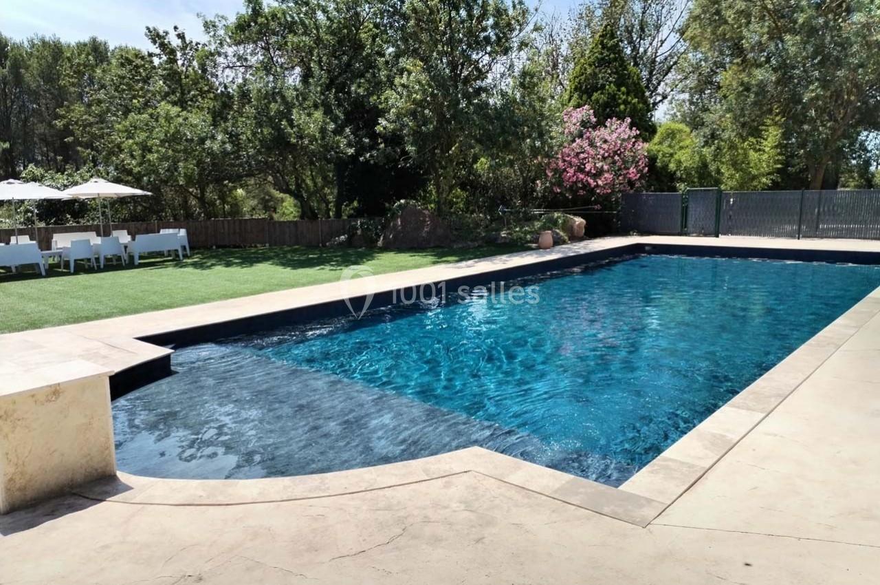 Piscine extérieure entourée de végétation, avec chaises longues et parasols sur une pelouse à l'arrière-plan.