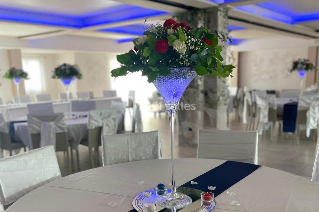 Centre de table floral avec roses rouges et feuillage, posé sur une table blanche dans une salle de réception décorée.