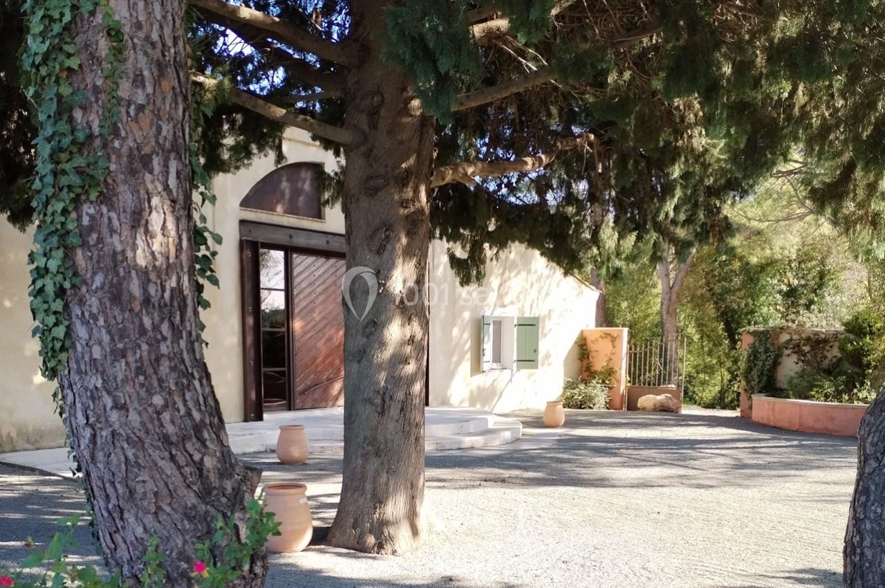 Façade d'une maison beige entourée d'arbres, avec une porte en bois et des pots en terre cuite sur une allée gravillonnée.