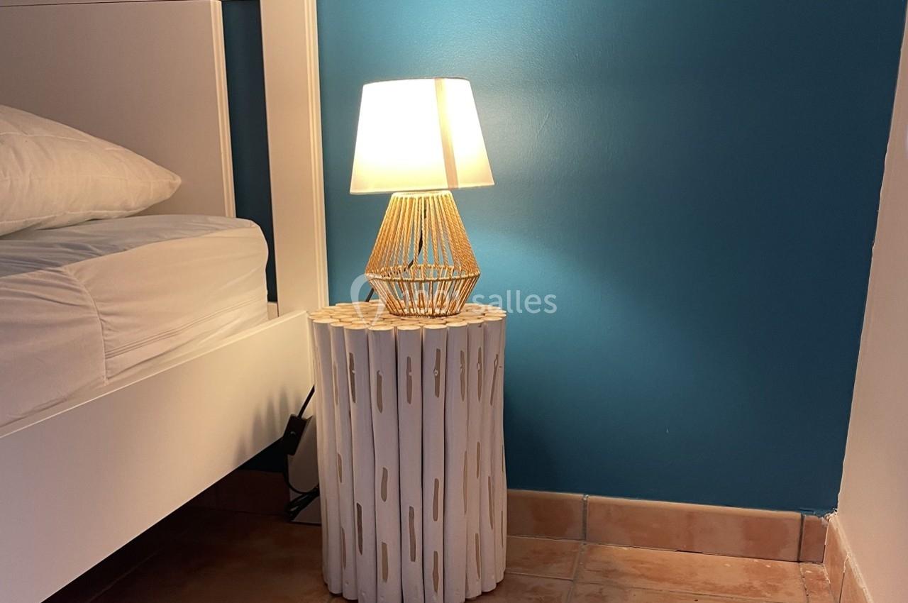 Lampe allumée sur une table d'appoint blanche en bois, placée à côté d'un lit avec un mur bleu en arrière-plan.