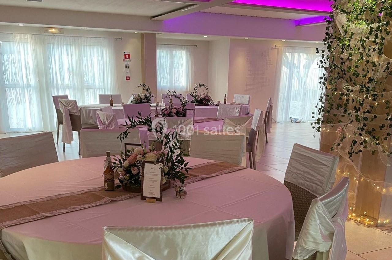 Salle de réception décorée avec des tables rondes, nappes blanches, chaises houssées et éclairage rose au plafond.