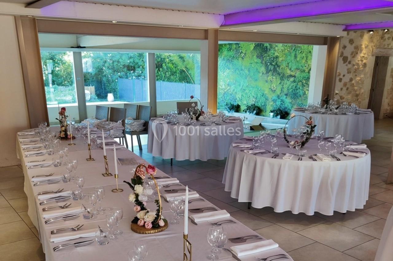 Salle de réception décorée avec des tables dressées, nappes blanches, chandeliers et vue sur un jardin verdoyant.