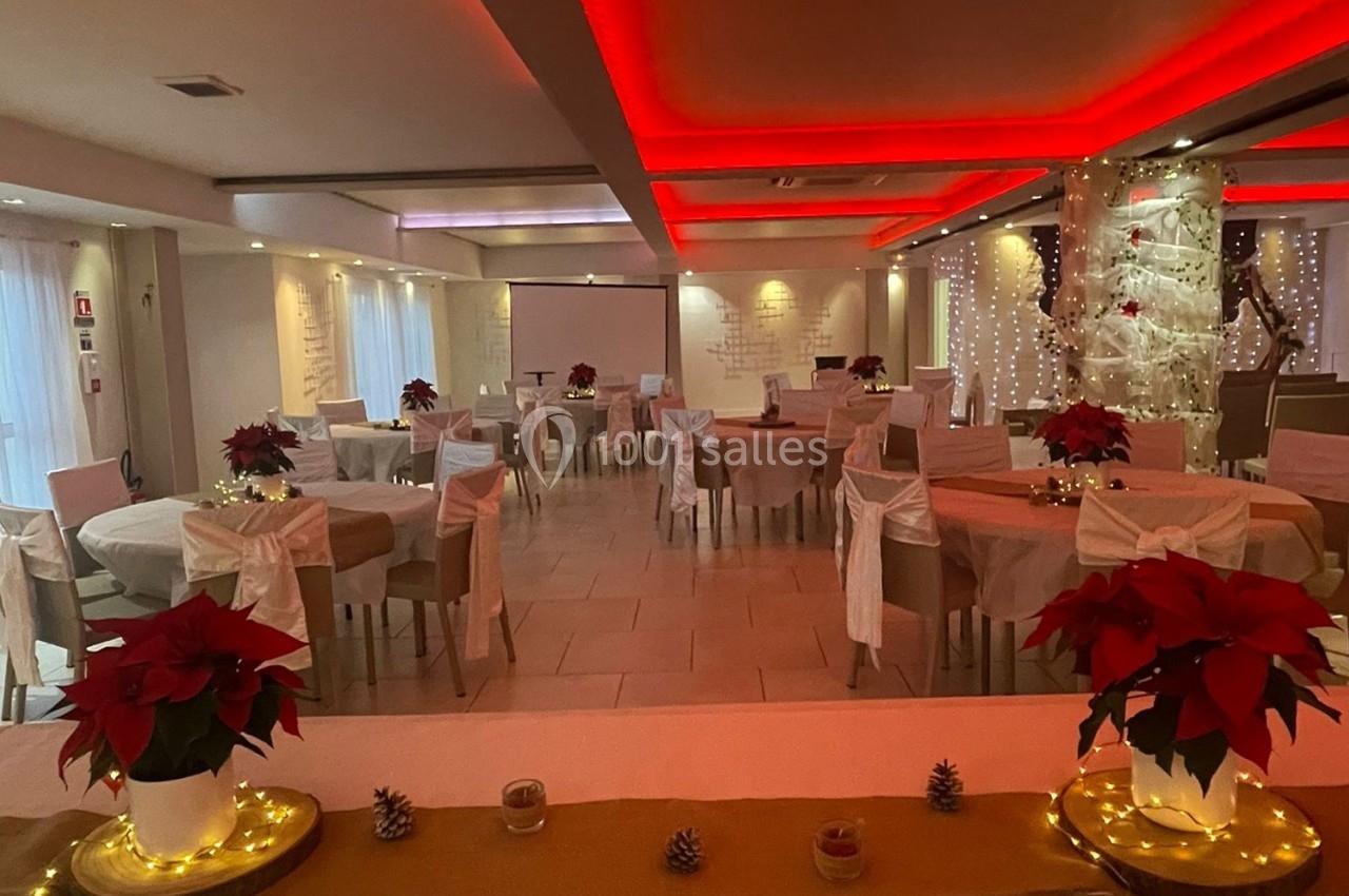 Salle de réception décorée pour une fête, avec tables rondes, nappes beige, chaises blanches et poinsettias rouges.