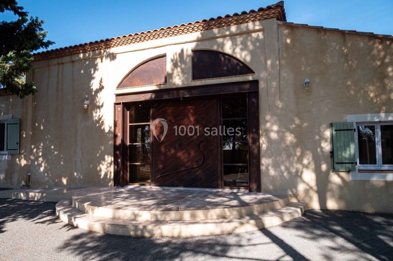 Façade d'un bâtiment en pierre avec une grande porte en bois sculptée, entourée d'ombres d'arbres.