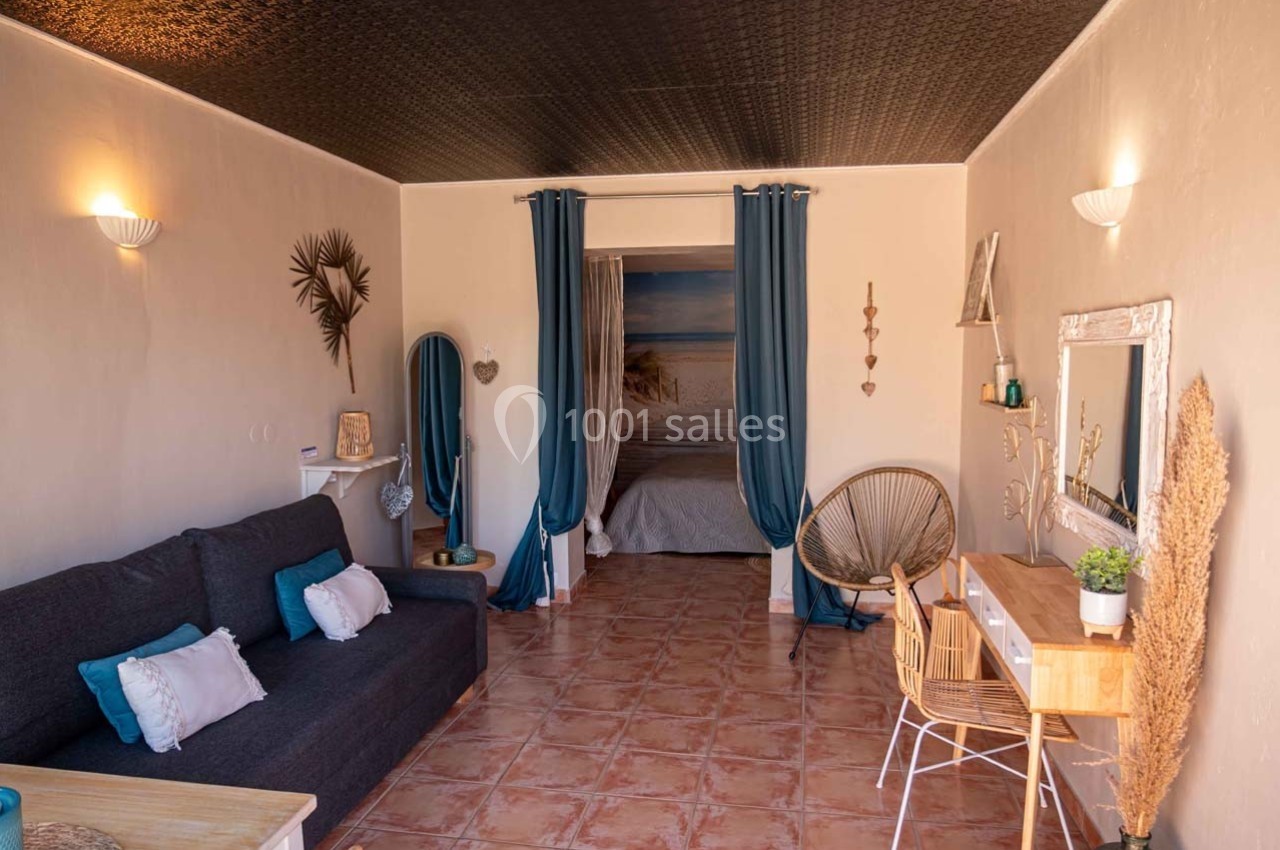 Salon lumineux avec canapé, bureau en bois, fauteuil en rotin et rideaux bleus encadrant une ouverture vers une chambre.