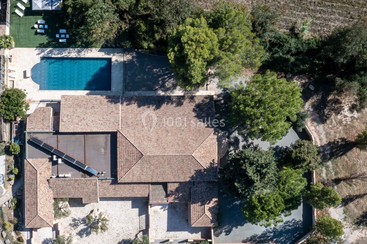 Vue aérienne d'une maison avec toit en tuiles, piscine, arbres environnants et terrain adjacent.