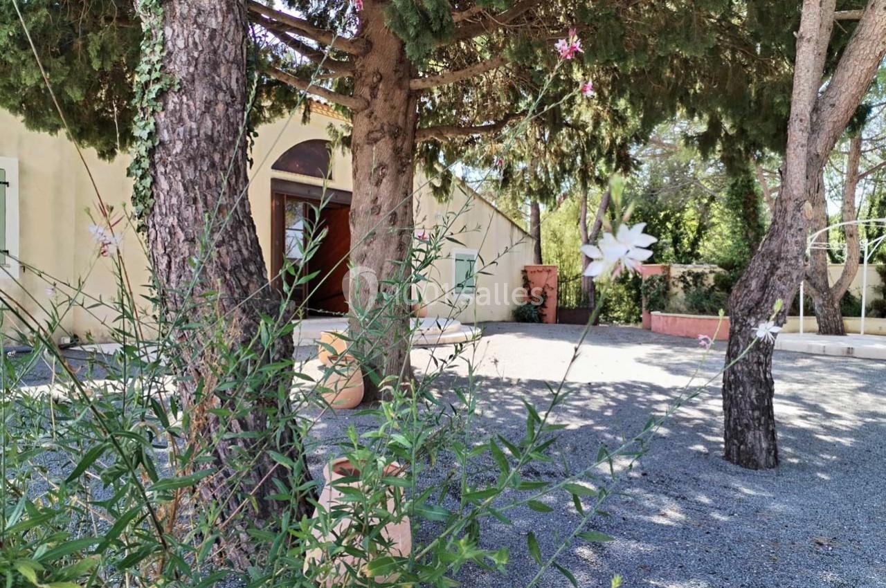 Vue d'une cour ombragée avec des arbres, des plantes fleuries et une façade beige en arrière-plan.