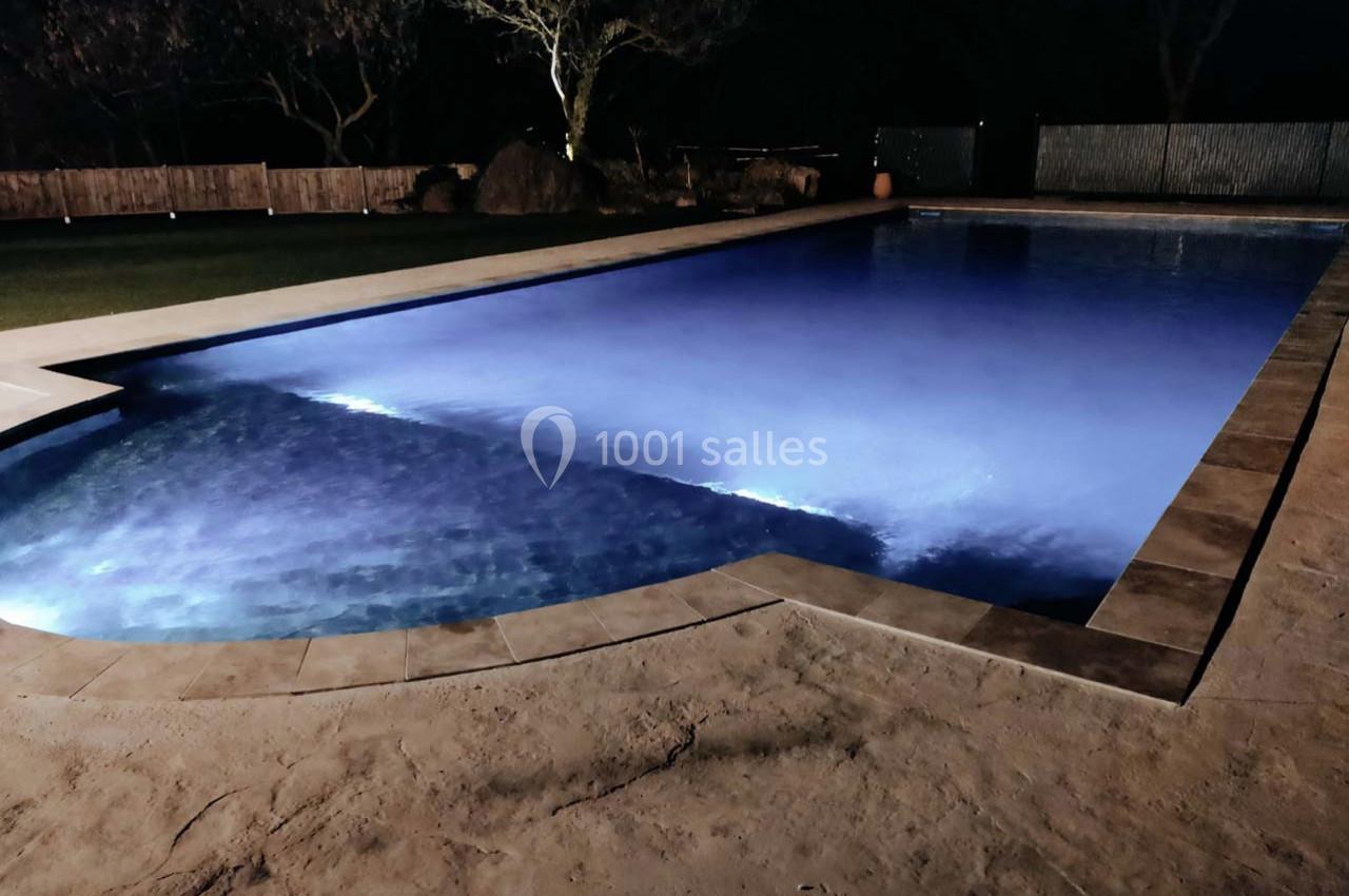 Piscine éclairée la nuit avec un éclairage bleu, entourée d'un sol en pierre et d'une clôture en bois.