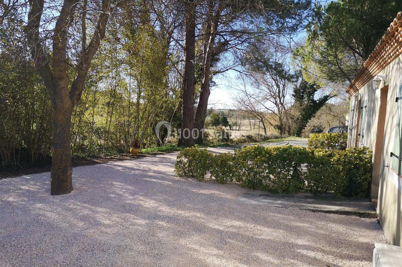 Cour extérieure en gravier bordée de haies et d'arbres, avec une maison à droite et un paysage rural en arrière-plan.