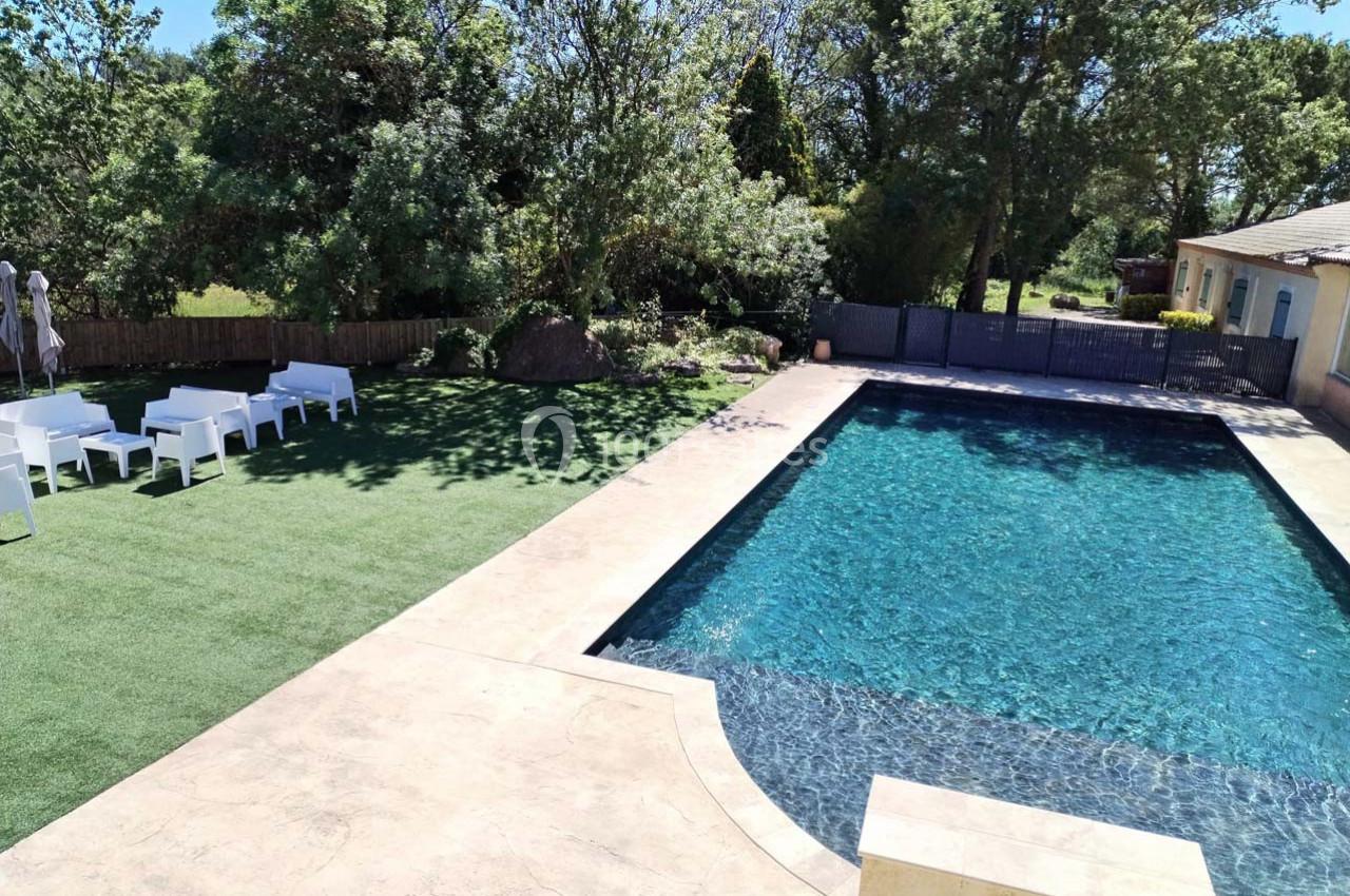 Piscine extérieure entourée de dalles claires, pelouse artificielle et mobilier de jardin blanc, avec arbres en arrière-plan.