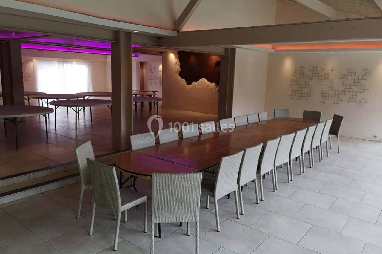Salle lumineuse avec une grande table en bois entourée de chaises, murs décorés et éclairage coloré en arrière-plan.