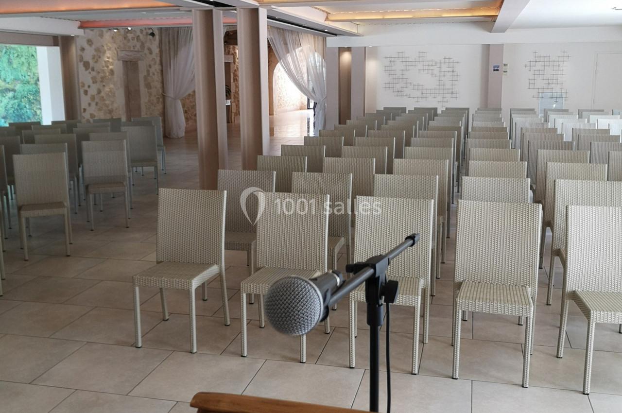 Salle de conférence lumineuse avec des rangées de chaises en osier beige et un micro sur un pupitre au premier plan.