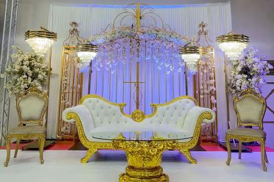 Salle de réception décorée avec des tables élégantes, des bouquets de fleurs et une estrade ornée d'un canapé doré.