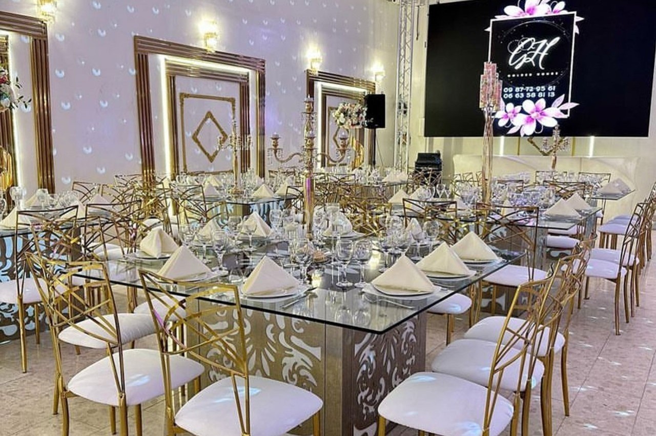 Salle de réception élégante avec tables en verre, chaises dorées, décor floral et éclairage tamisé.