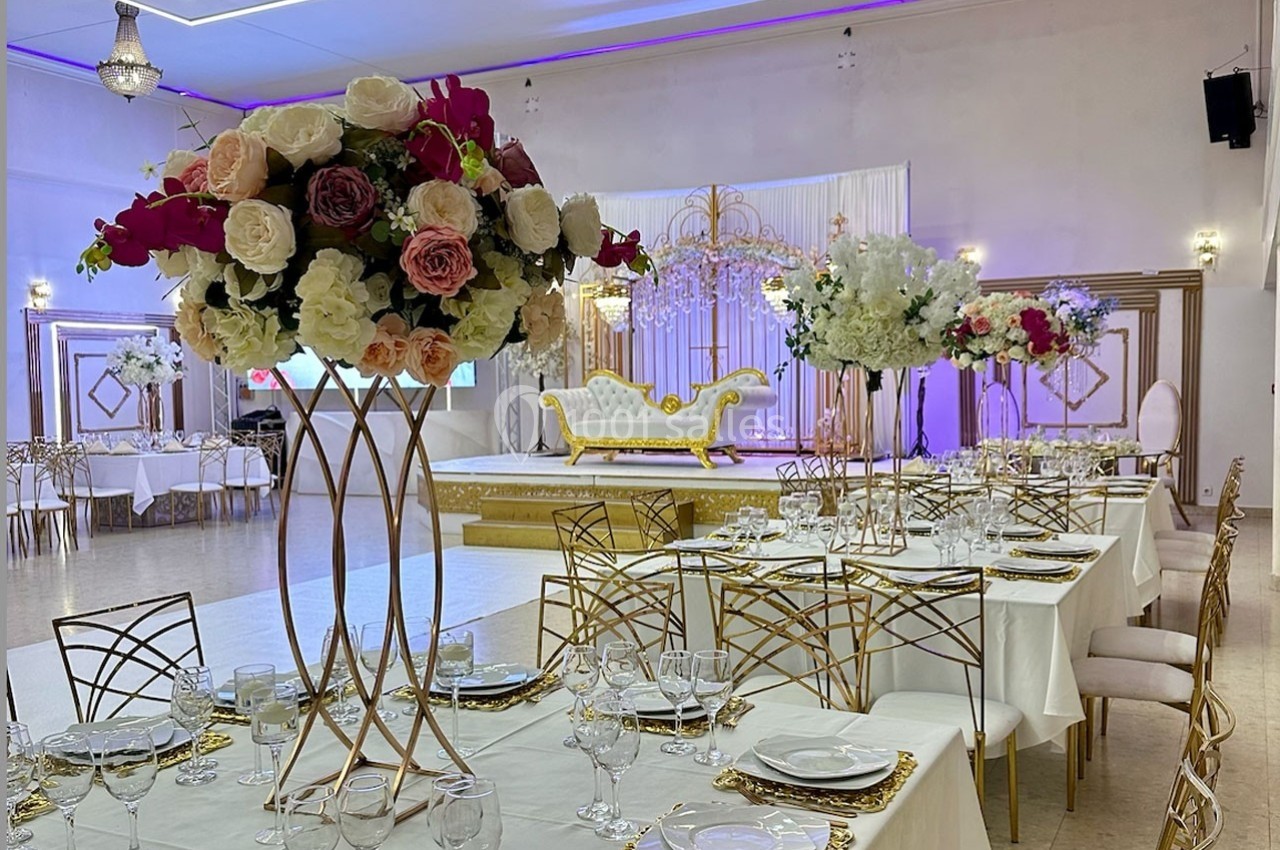 Salle de réception décorée avec des tables dressées, des centres de table fleuris et une scène ornée d'un canapé doré.