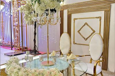 Salle de réception décorée avec des tables élégantes, des bouquets de fleurs et une estrade ornée d'un canapé doré.