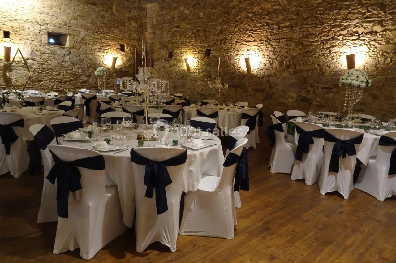 Salle de réception avec murs en pierre, tables rondes dressées, nappes blanches et chaises décorées de nœuds noirs.