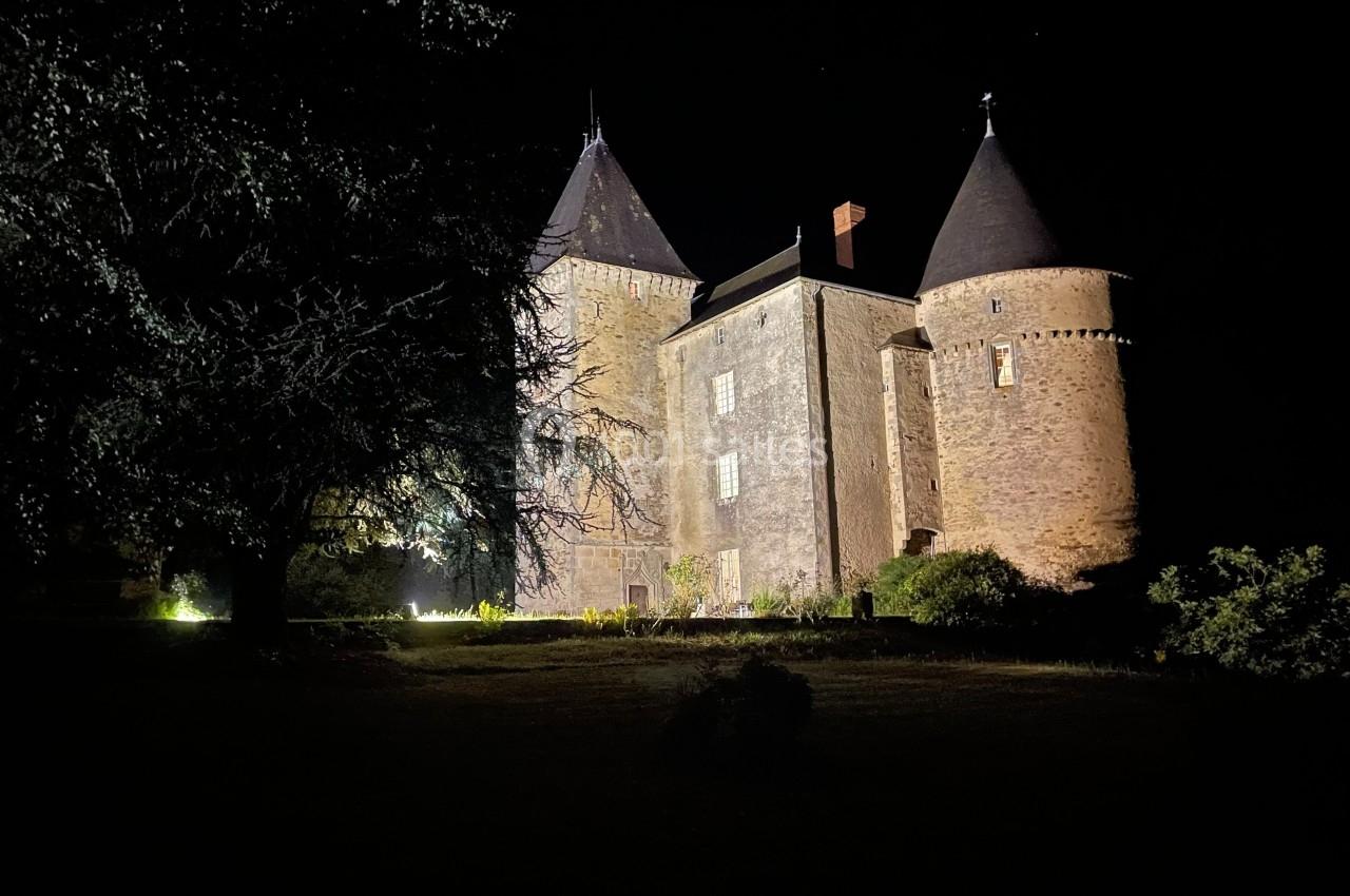 Château en pierre éclairé de nuit, entouré d'arbres et de végétation dans un cadre sombre.