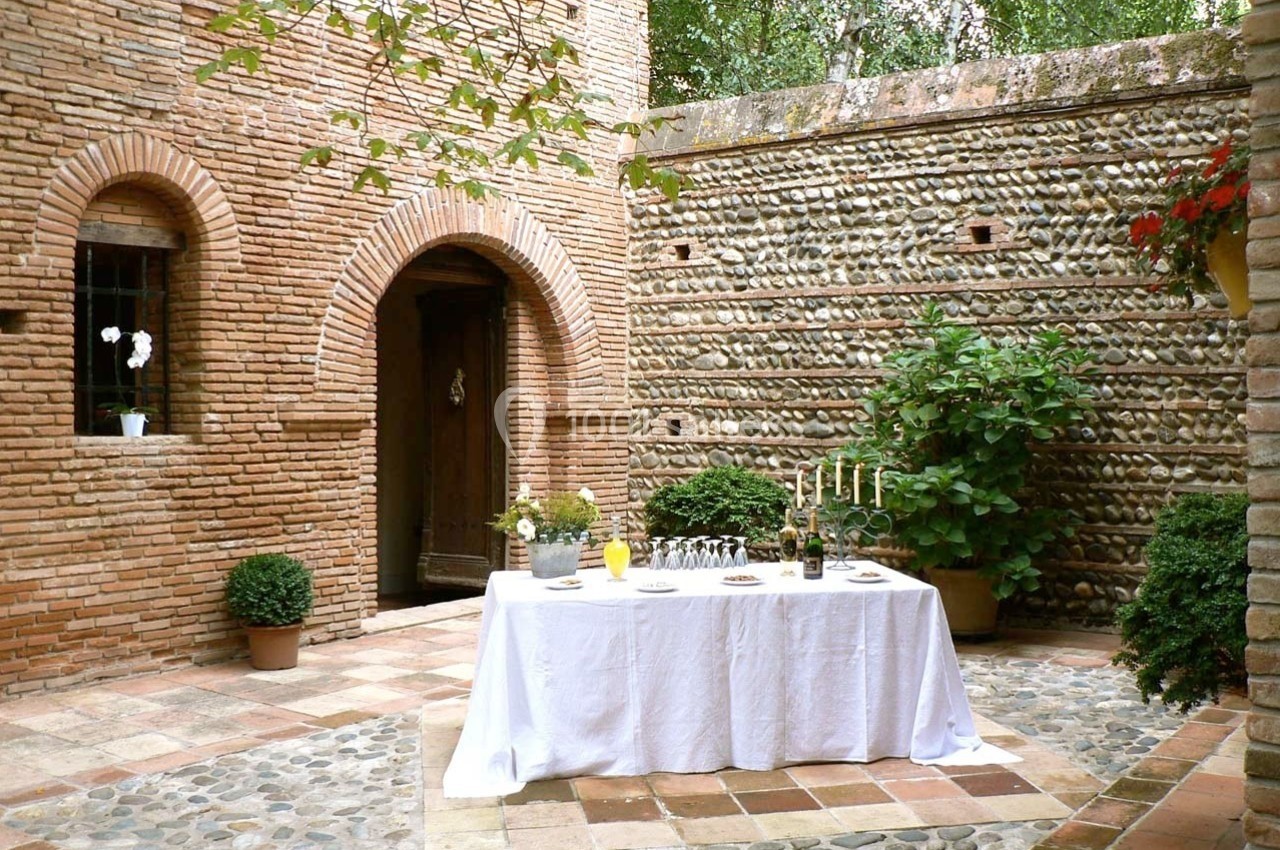Table dressée avec nappes blanches dans une cour en briques et galets, entourée de plantes et d'une porte en bois.