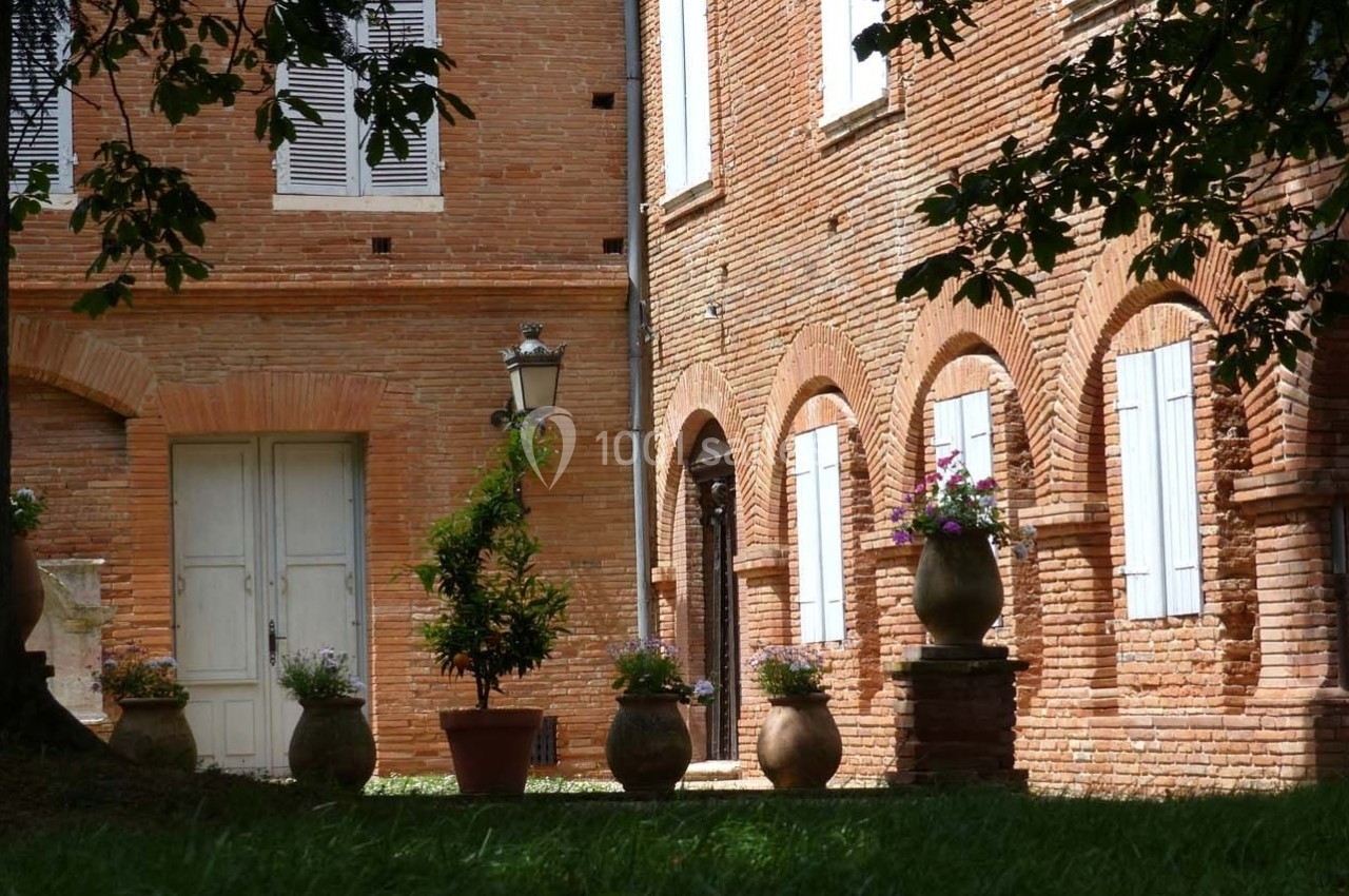 Cour bordée de murs en briques rouges avec des arches, des fenêtres à volets blancs et des pots de fleurs alignés.