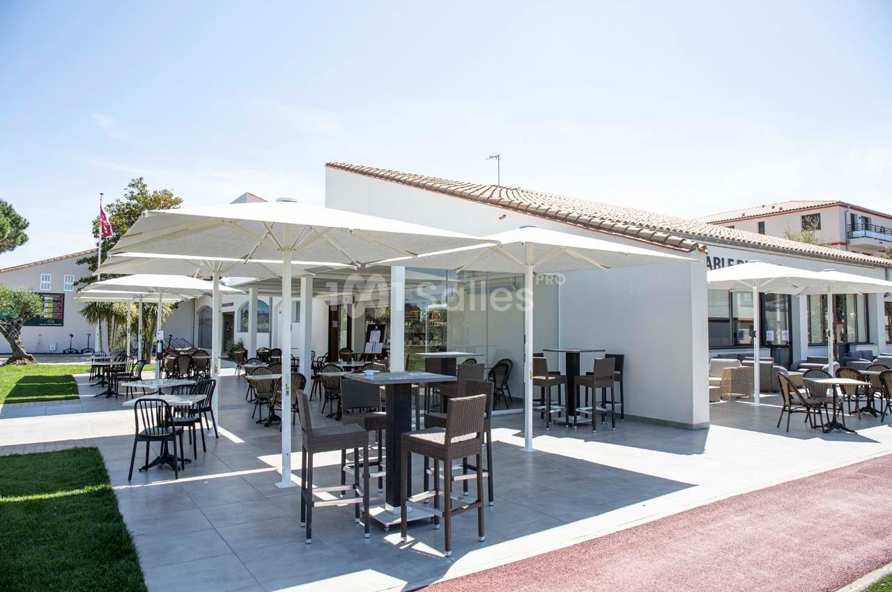 Terrasse extérieure avec tables, chaises et parasols, entourée de pelouse et bâtiments modernes en arrière-plan.
