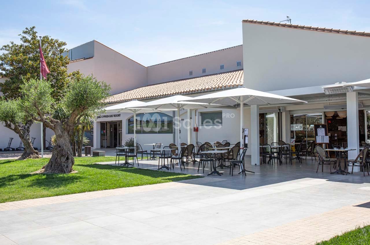 Terrasse extérieure d'un bâtiment moderne avec tables, chaises, parasols et un olivier sur une pelouse.