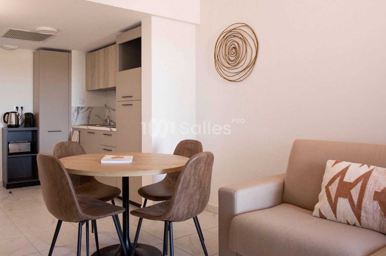 Salle à manger avec table ronde, quatre chaises, canapé beige, cuisine ouverte et décoration murale en spirale.