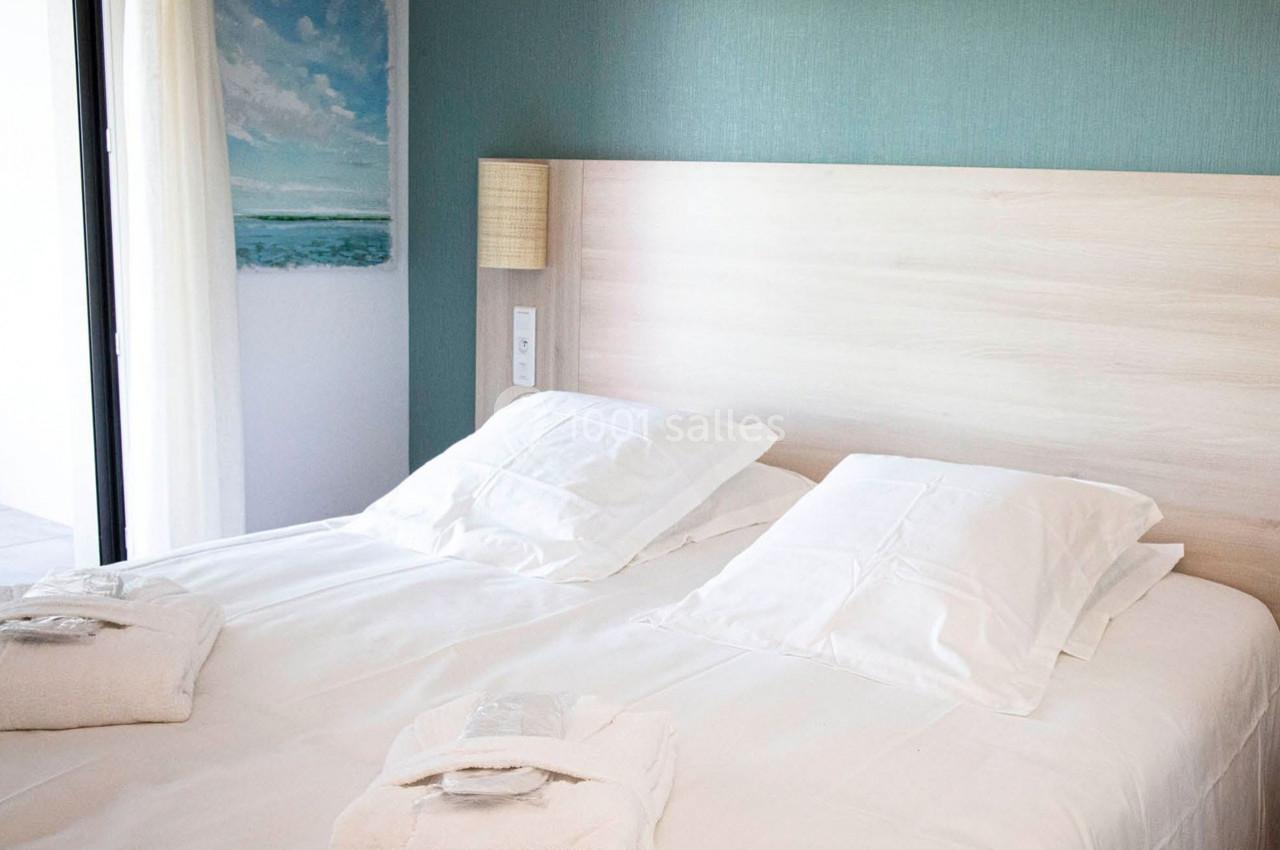 Lit double avec draps blancs, deux oreillers et peignoirs pliés, dans une chambre lumineuse au mur vert pastel.