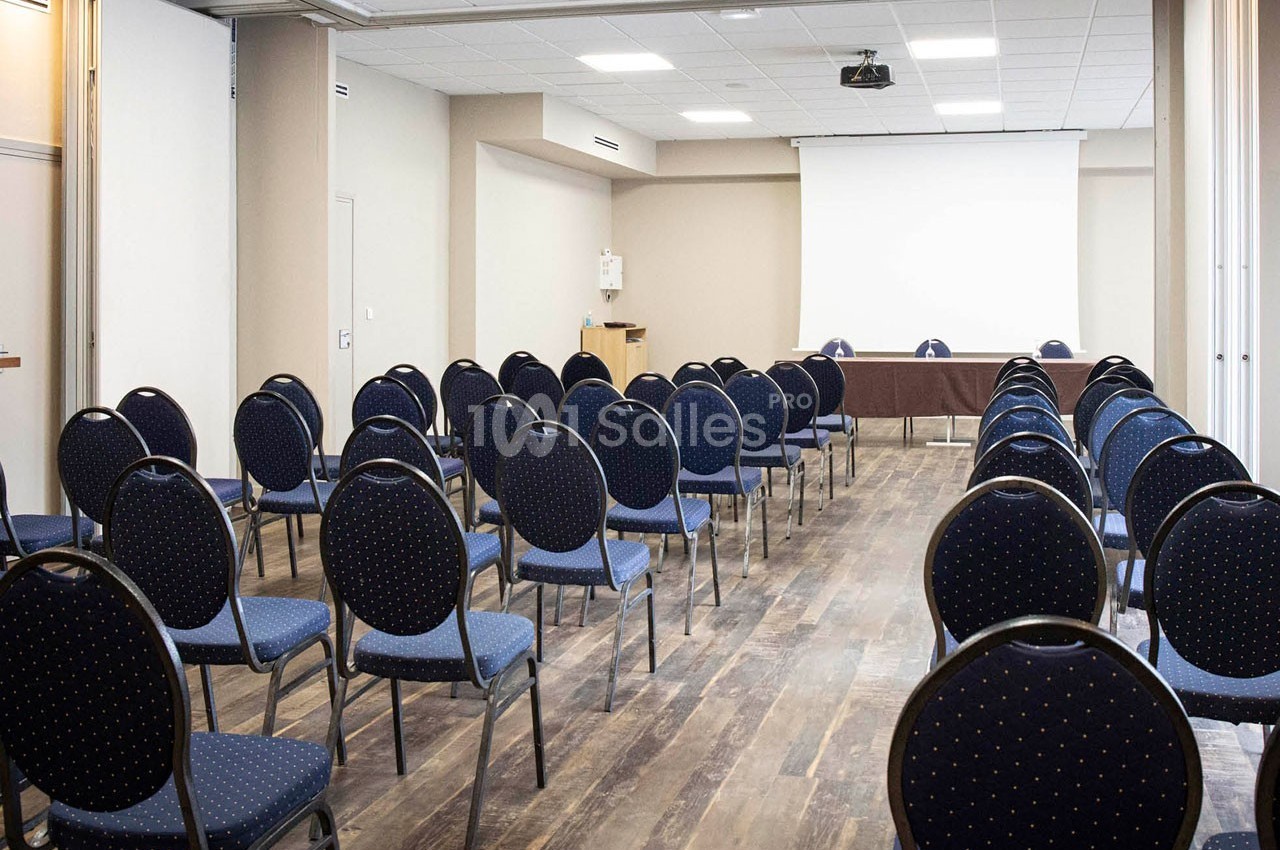 Salle de conférence vide avec des rangées de chaises bleues et un écran de projection blanc à l'avant.