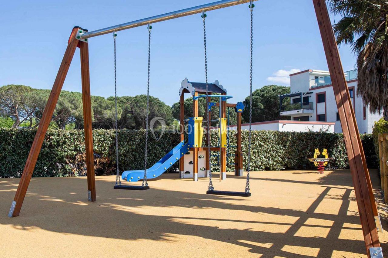 Structure de jeux pour enfants avec toboggan et balançoires, installée sur un sol sablé entouré de végétation.