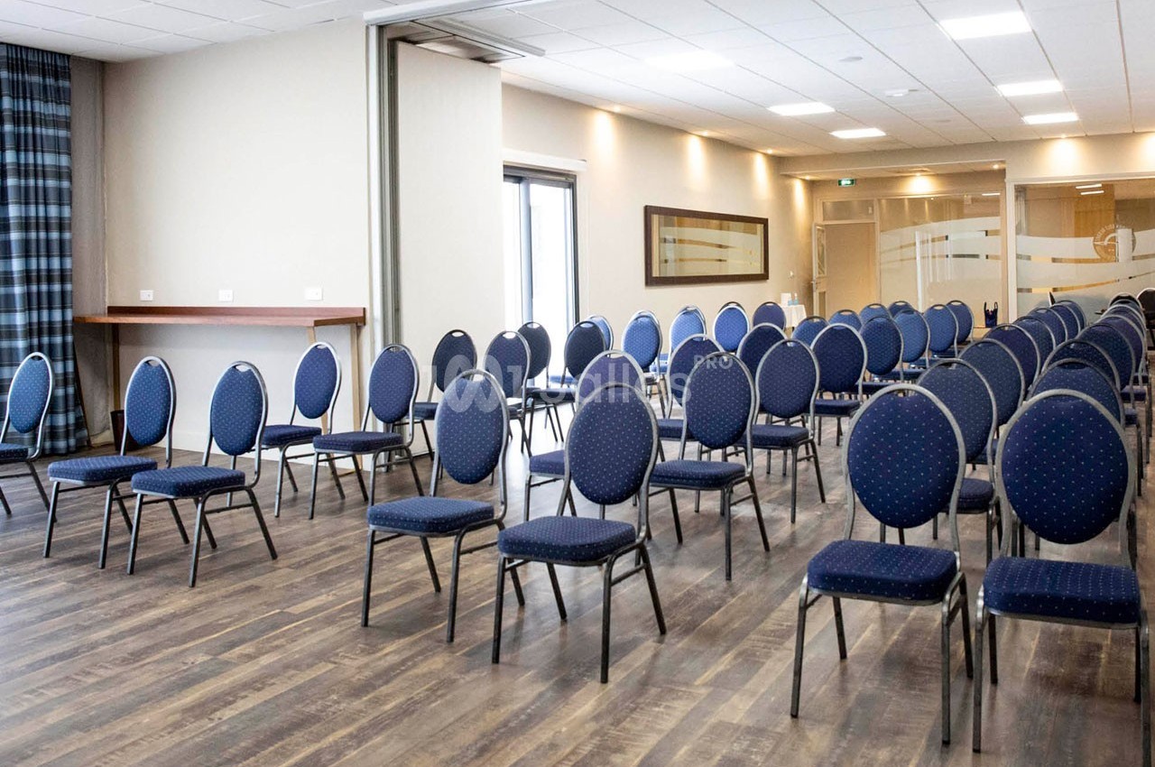 Salle de réunion avec des chaises bleues disposées en rangées sur un sol en bois, éclairée par des lumières au plafond.