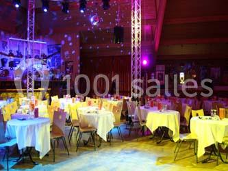 Salle de réception éclairée avec des tables rondes dressées, des chaises et une scène en arrière-plan.