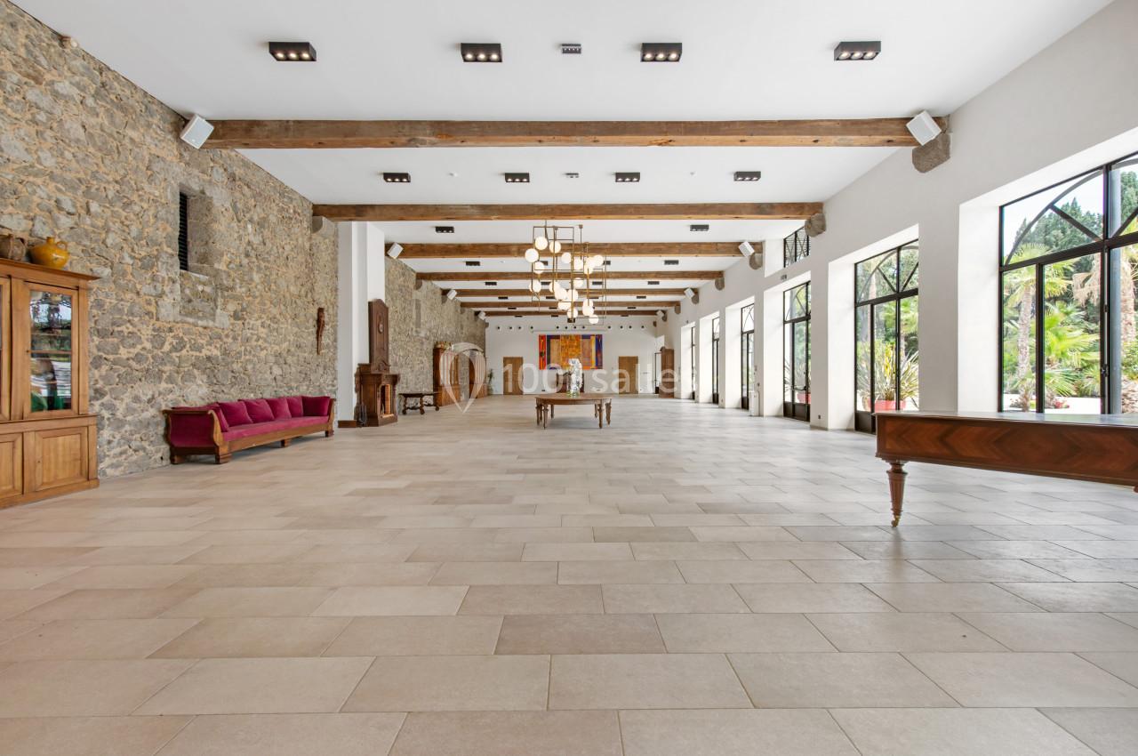 Salle spacieuse avec murs en pierre, grandes fenêtres, poutres apparentes et mobilier en bois.