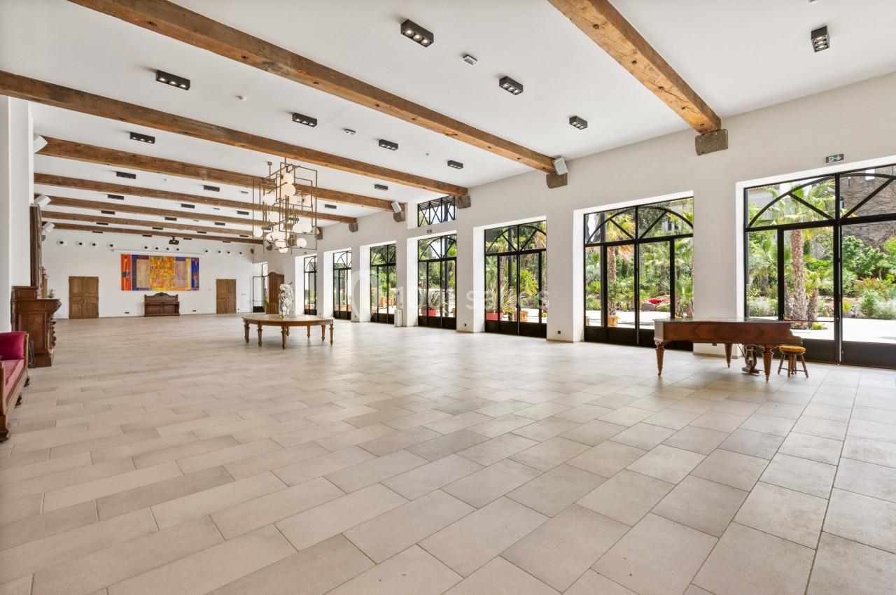 Grande salle lumineuse avec sol en carrelage clair, poutres apparentes et larges fenêtres donnant sur un jardin verdoyant.