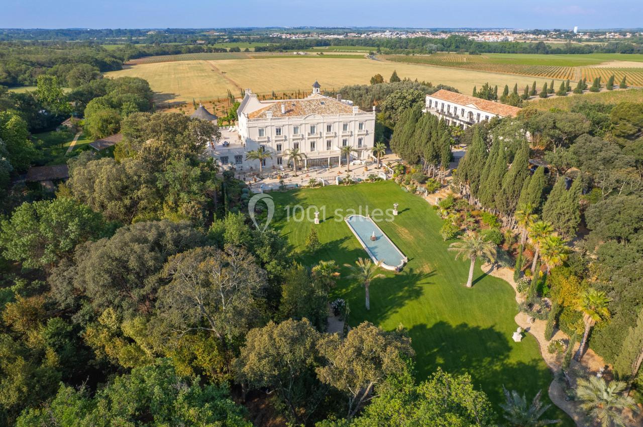 Vue aérienne d'un domaine avec un grand jardin, une fontaine centrale et une bâtisse entourée de végétation et de champs.