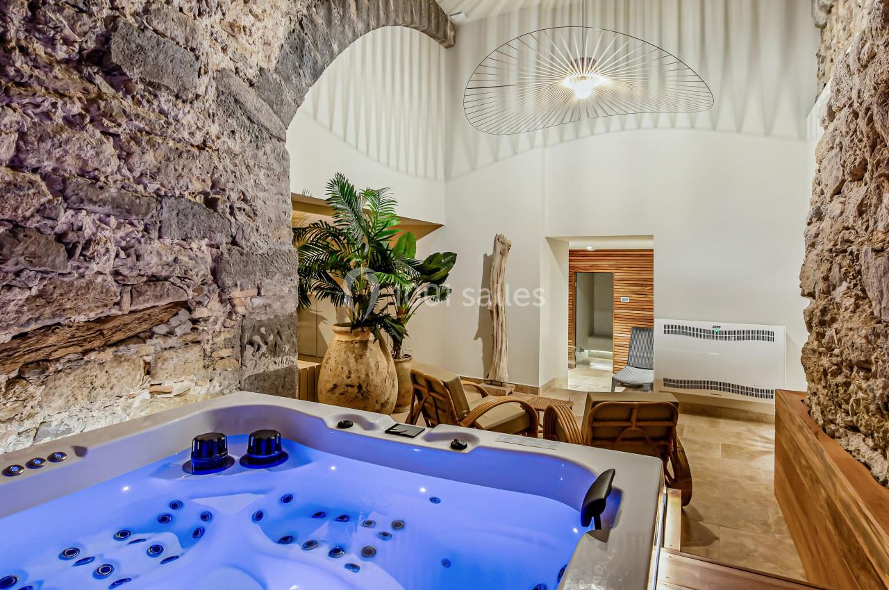 Jacuzzi éclairé dans un espace intérieur avec murs en pierre, plantes décoratives et chaises longues.