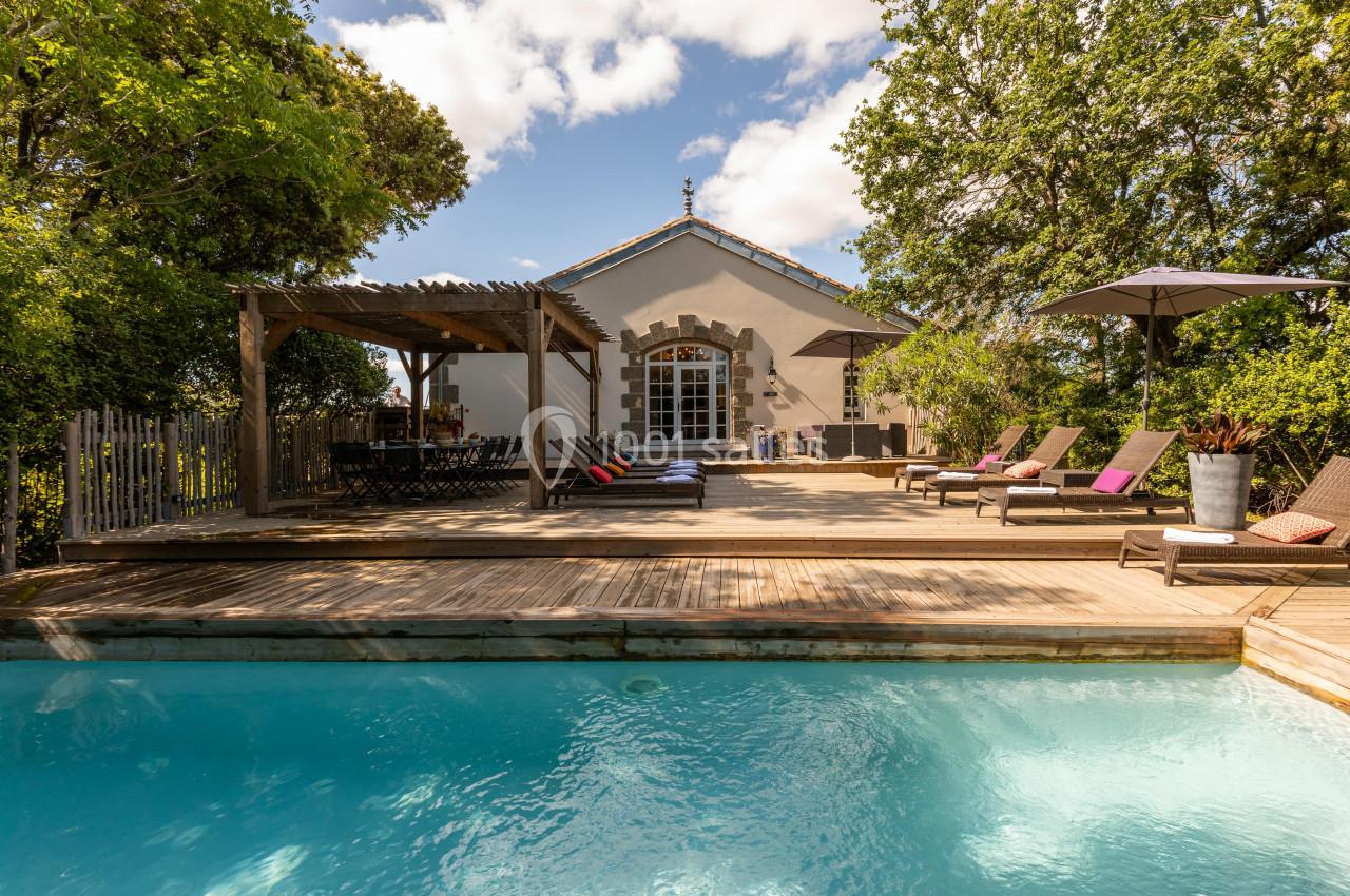 Terrasse en bois avec piscine, transats, pergola et maison en arrière-plan entourée de végétation.