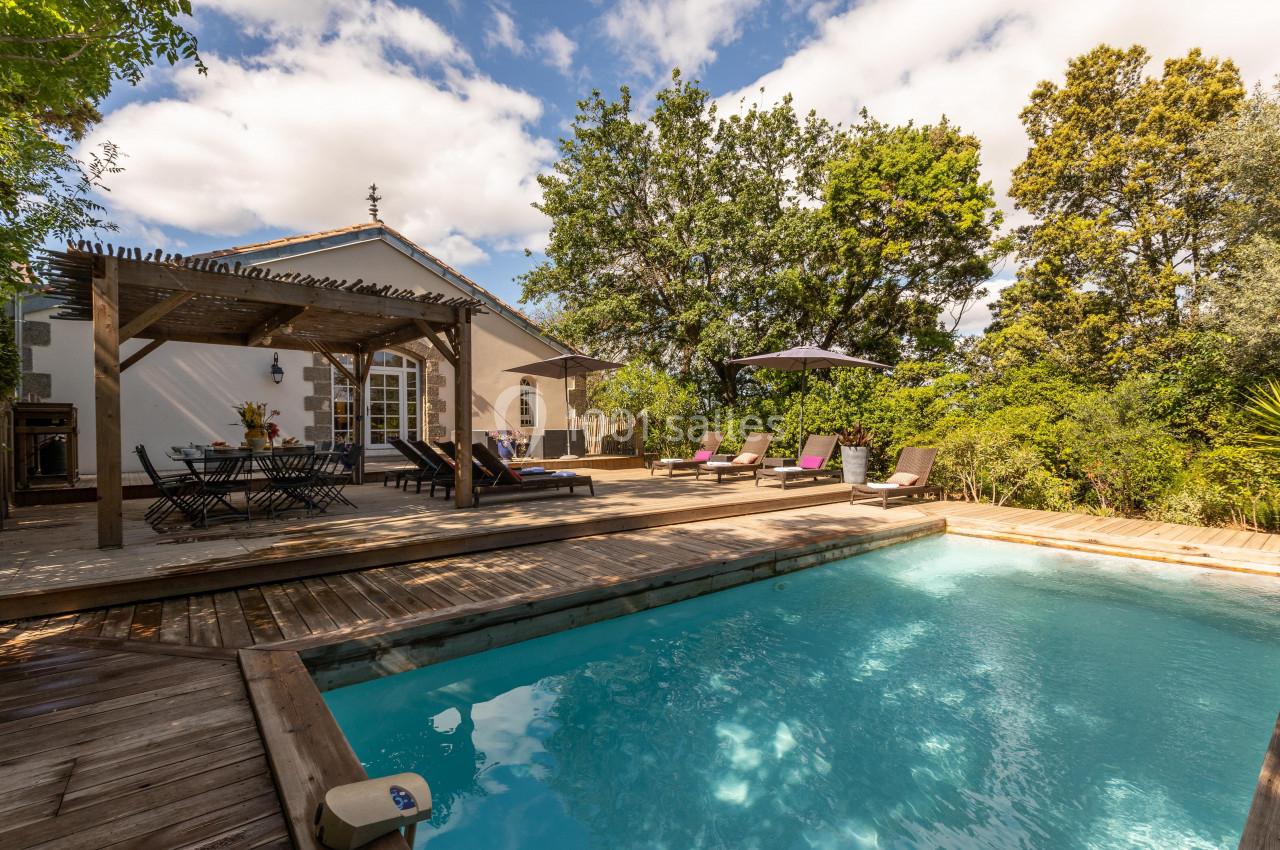 Piscine extérieure entourée de transats et d'une terrasse en bois, avec une pergola et une maison en arrière-plan.