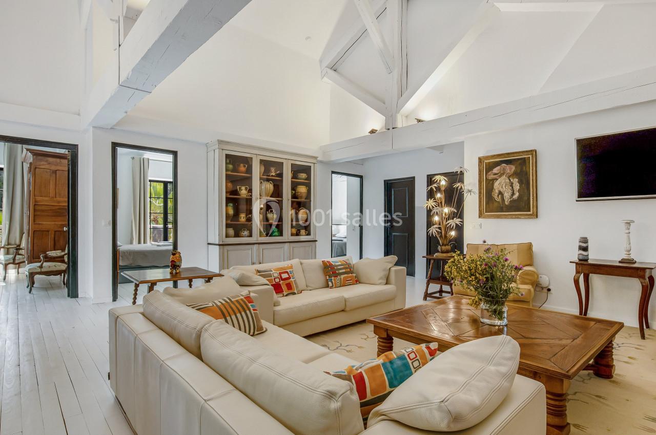 Salon lumineux avec canapé d'angle blanc, table basse en bois, étagères vitrées et poutres apparentes blanches.