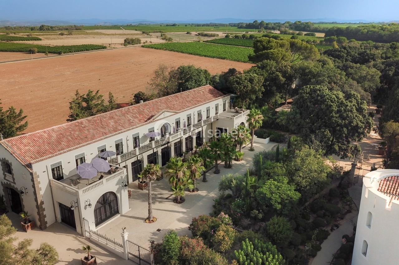Vue aérienne d'un bâtiment de style méditerranéen entouré de jardins, avec des champs et des collines à l'arrière-plan.