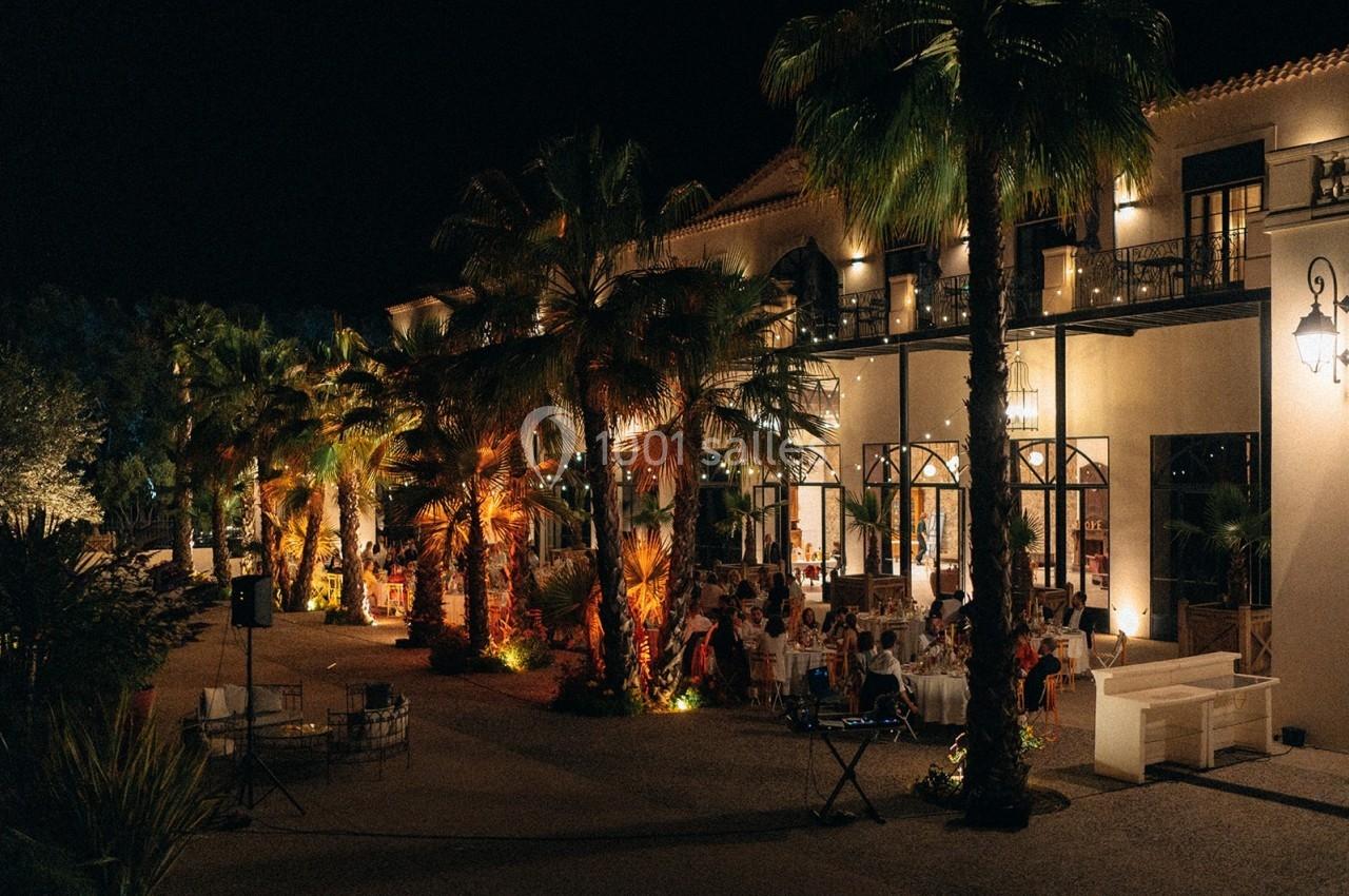 Terrasse éclairée d'un bâtiment avec des palmiers et des tables occupées par des convives en soirée.