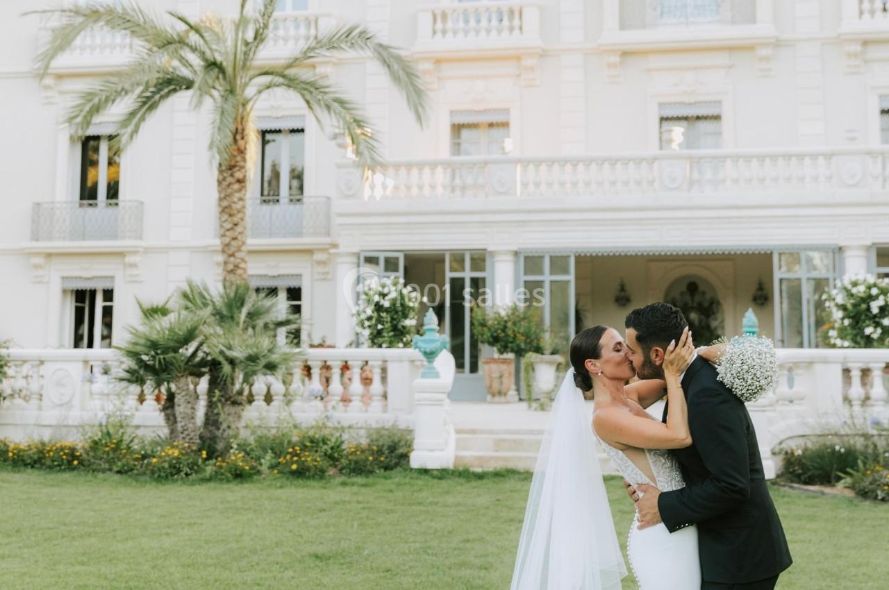 Un couple de mariés s'embrasse sur une pelouse devant un bâtiment blanc élégant avec des palmiers.