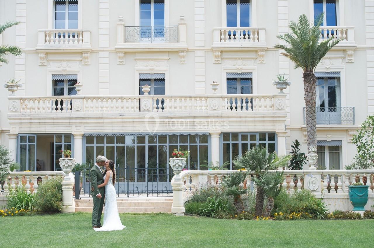 Un couple en tenue de mariage se tient devant un bâtiment élégant avec une façade beige et un jardin verdoyant.