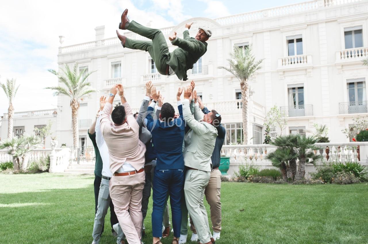 Un groupe d'hommes en tenue formelle lance un homme en costume vert en l'air devant un bâtiment élégant.