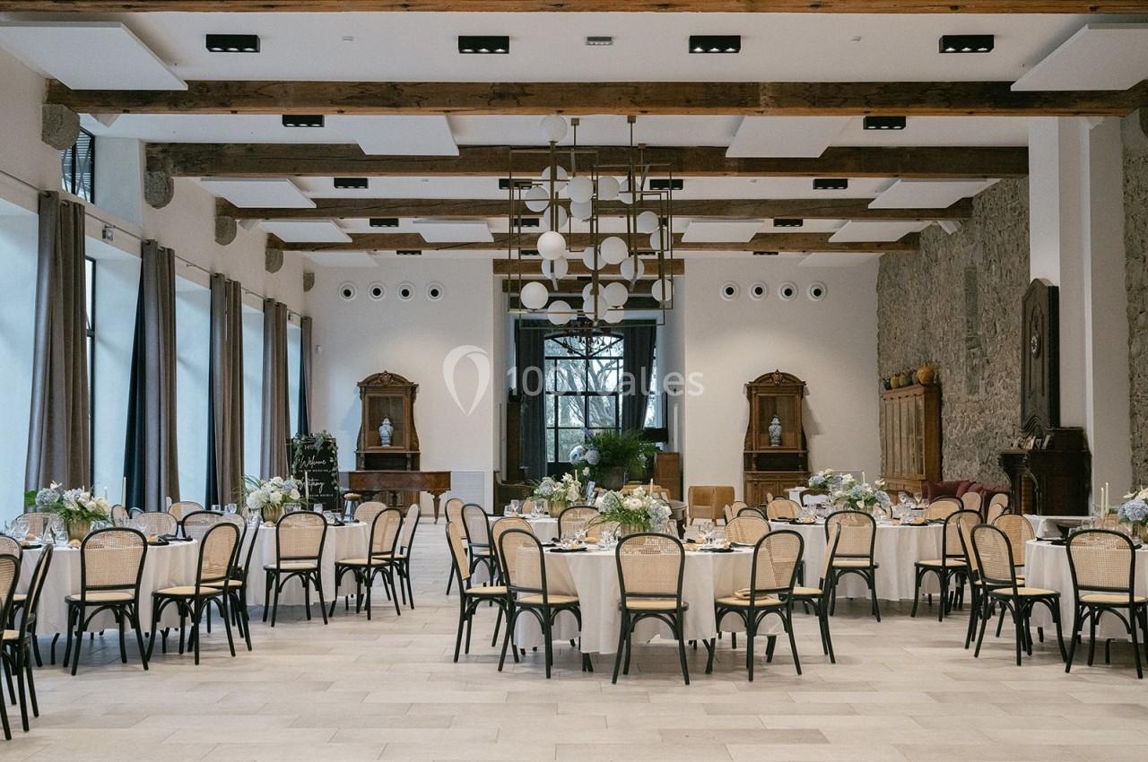 Salle de réception lumineuse avec tables rondes dressées, chaises en bois clair et poutres apparentes au plafond.