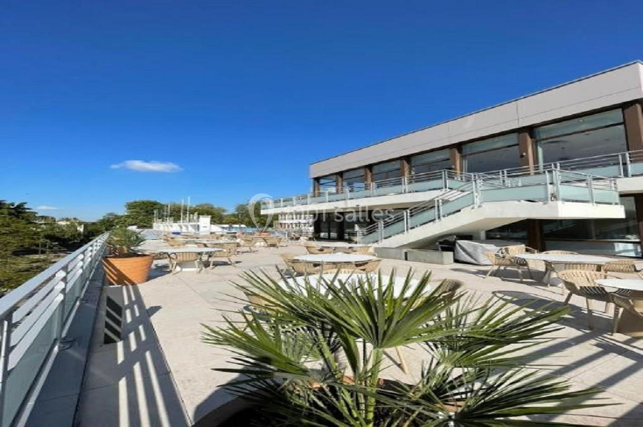 Location salle Nogent-sur-Marne (Val-de-Marne) - Hôtel du Port #1 Terrasse ensoleillée avec tables et chaises, bordée de plantes, devant un bâtiment moderne sous un ciel bleu.