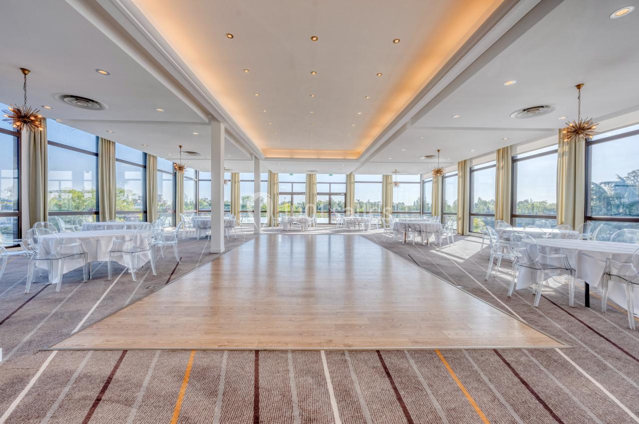 Salon Panoramique Salle lumineuse avec de grandes baies vitrées, tables rondes dressées et parquet central entouré de moquette.