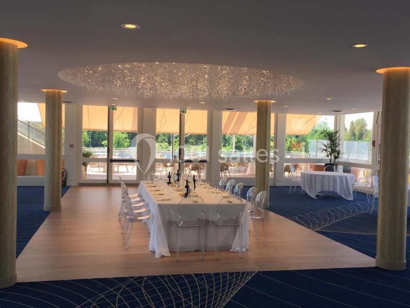 Salon Plaisance Salle lumineuse avec une grande table dressée, chaises transparentes, lustre moderne et baies vitrées donnant sur l…