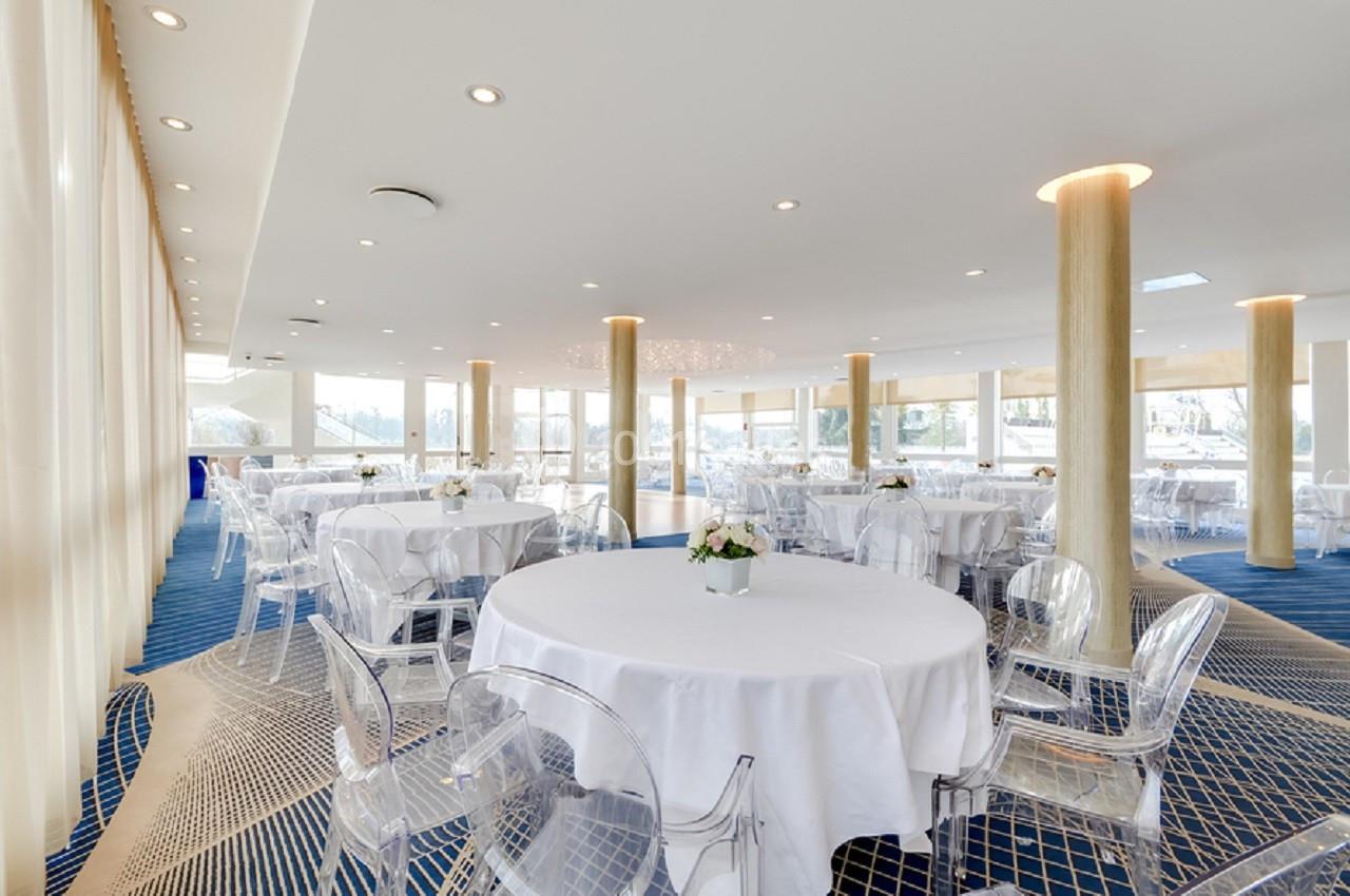 Salon Plaisance Salle lumineuse avec grandes baies vitrées, tables rondes dressées de nappes blanches et chaises transparentes.