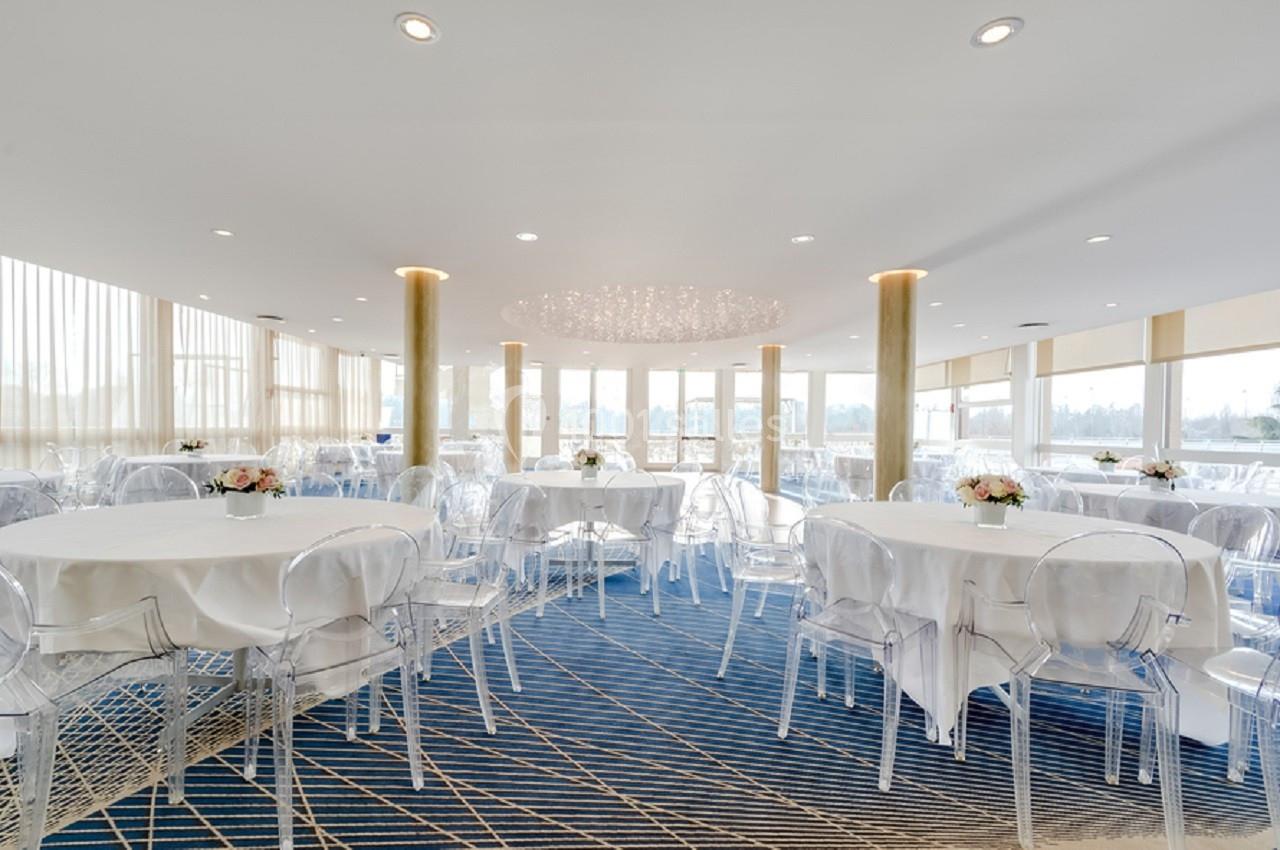 Salon Plaisance Salle lumineuse avec tables rondes dressées, chaises transparentes et grandes baies vitrées donnant sur l'extérieur.